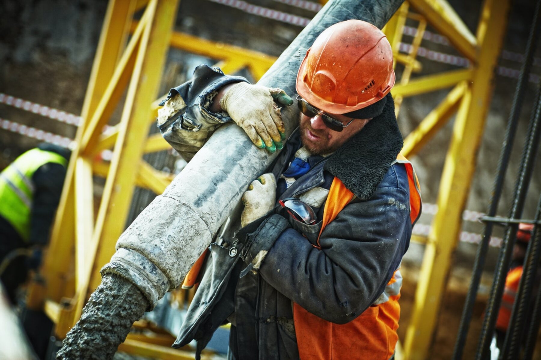 A worker in a hard hat and high-visibility jacket carrying pipe across yellow structural steel