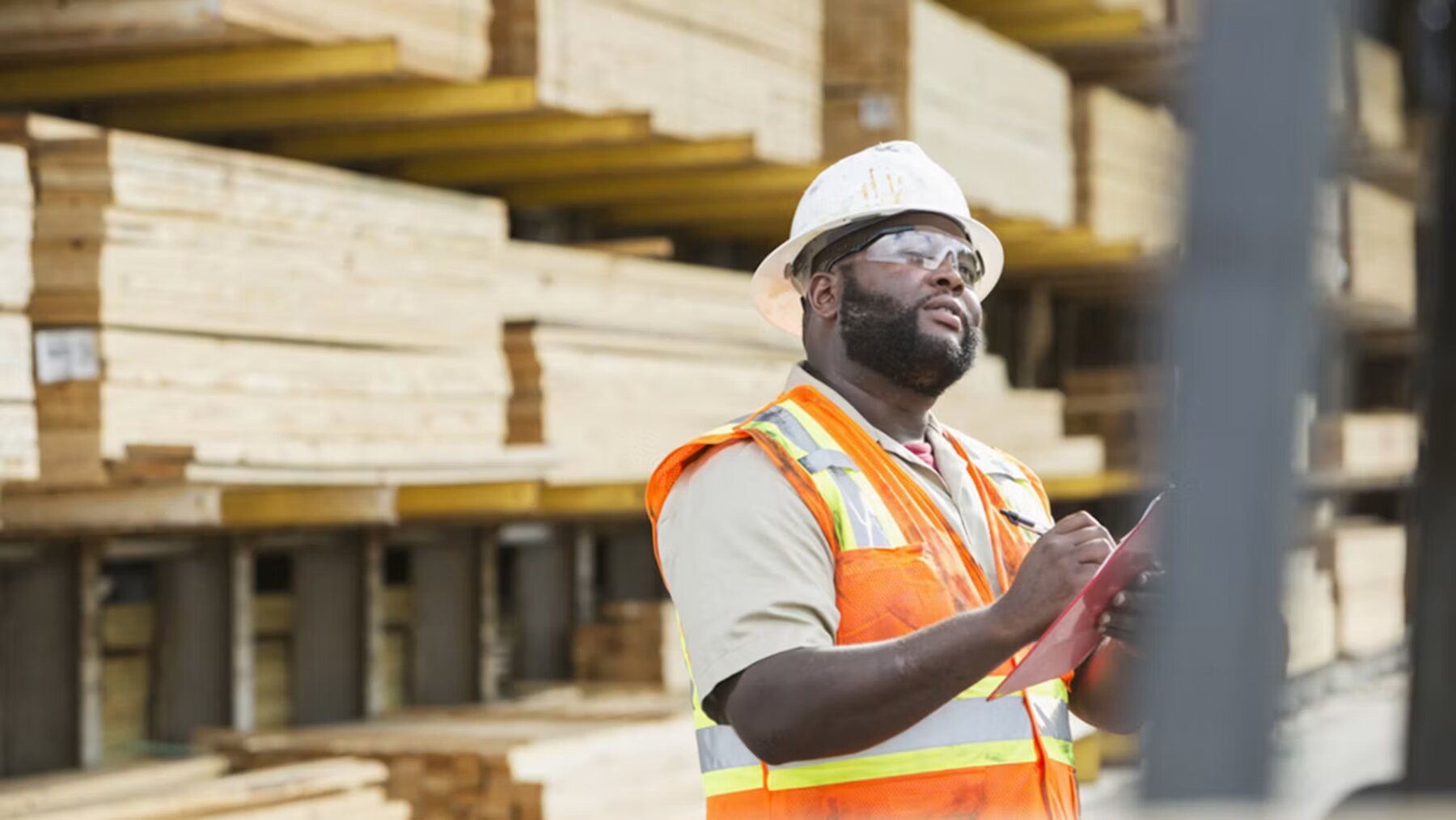 A supervisor in a hard hat and safety glasses checking a list in front of stacked timber