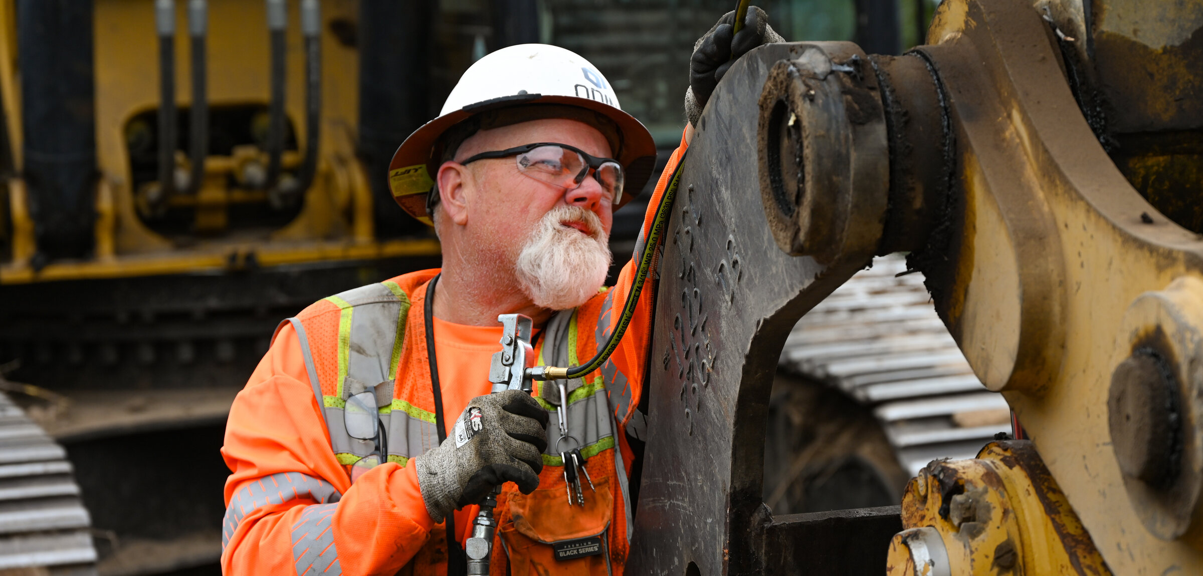An equipment technician in a hard hat and high-visibility gear greasing the boom of a tracked excavator